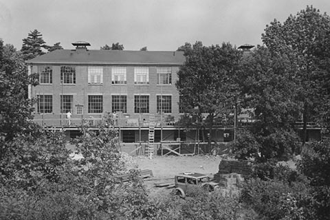 Photo shows the construction of Sydeman Hall underway, with workers on scaffolding, a ladder, and piles of construction materials in the foreground.