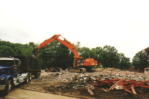 Behind a pile of rubbe, an orange excavator loading debris into a truck to be hauled away.