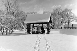 Two sets of footprints in the snow leading to the Old Wishing Well where two people are standing, 1952.