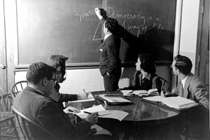 Max Learner writing on a blackboard while 4 students, seated at a round table, look on. December 16, 1949. 