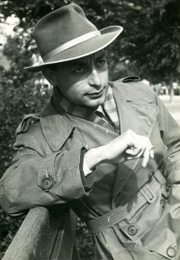 Young man in trench coat and hat, smoking a cigarette, sitting on a bench