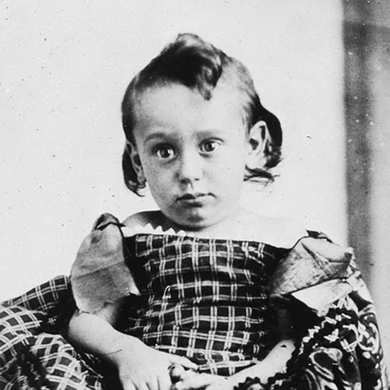 A 2 year old Louis Brandeis seated with tall white socks, lace up shoes, and a large fabric covering (black and white photograph)