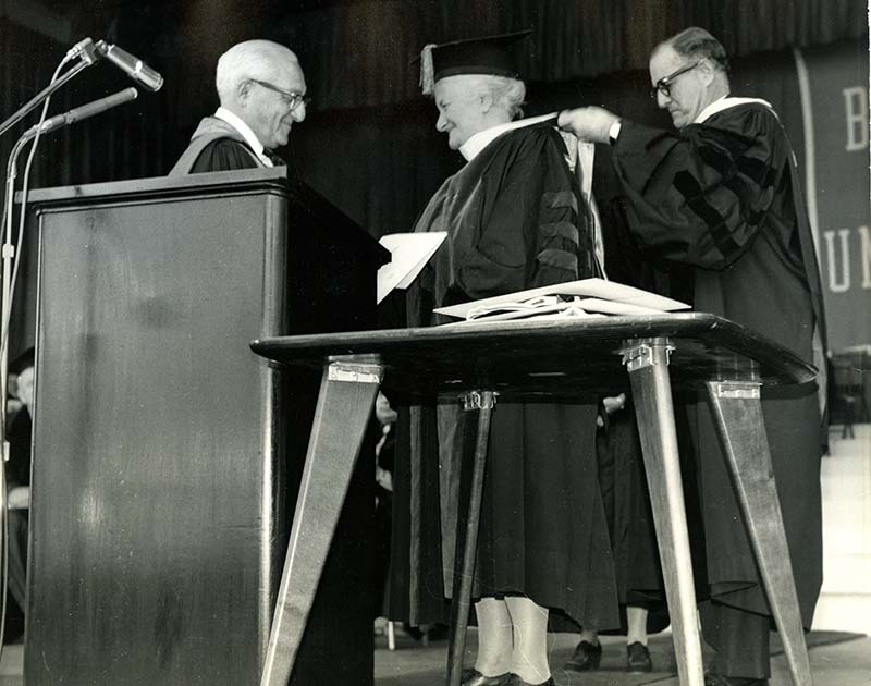 Susan Brandeis receiving her Honorary Degree from Abram Sachar at Brandeis University's 1963 Commencement, while the degree hood is placed around her neck (black and photograph)