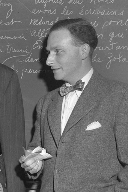 Irving Fine, side view, wearing a bowtie and tweed jacket, standing in front of a blackboard with French writing.