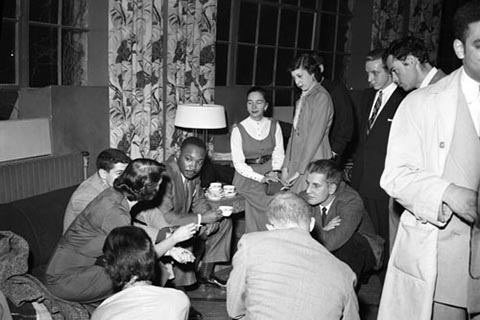 Dr. Martin Luther King, Jr. and Brandeis students engaged in conversation at a reception following his speech. Some students sit with him on a sofa holding teacups, while others gather around, standing.