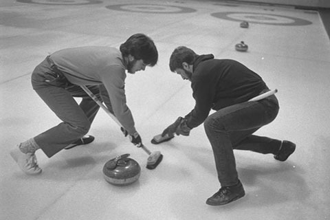 Two men are competing in a curling match. They are sweeping the ice in front of the curling stone with their curling brooms.