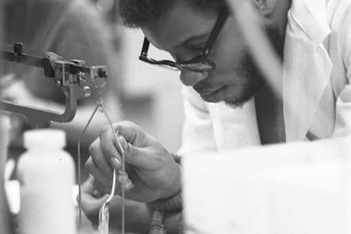 A man wearing glasses and a lab coat is leaning over a table. He's holding a spoon and pouring a substance into a container. There are scientific instruments around him.