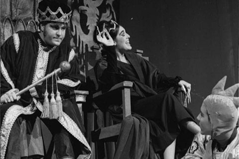 A male performer dressed as a king and a female performer dressed as a queen are sitting on two thrones. The women is talking and gesturing her hand while the man looks frustrated. A performer is sitting on the steps below them and is looking up at them.