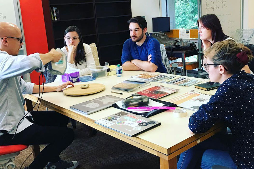 Students sit around a table and watch an instructor use equipment in the Digital Scholarship Lab