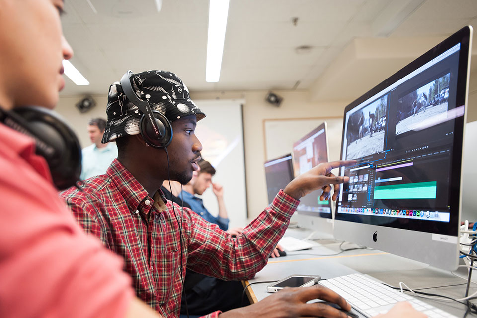 A student wearing headphones is pointing to a computer screen where he is editing a video