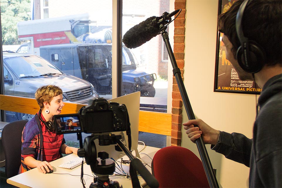 Person sitting at computer being recorded on video while someone else holds a microphone overhead