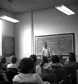 Teacher leaning on desk in front of class with a blackboard behind him filled with names of filmmakers.