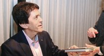 Photo of Mitch Albom, seated at a table, as he hands a book to a woman.