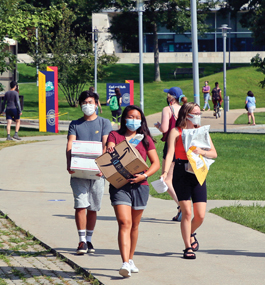 Three students wearing masks walk on a campus sidewalk carrying packages.