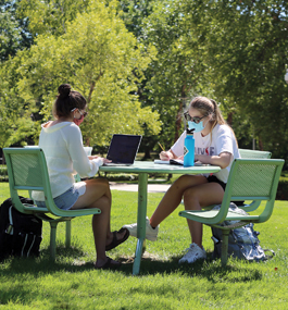 Two students sit at a round table on campus grounds surrounded by laptop, books and book bags