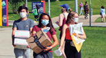 Three students wearing masks walk on a campus sidewalk carrying packages.