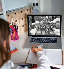 Photo of a student at a desk, looking at a laptop showing an image of Dr. Martin Luther King Jr. delivering his "I Have a Dream" speech