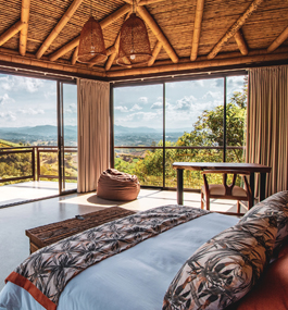Photo of a bedroom with a beamed ceiling and three large floor-to-ceiling windows that look out on a private porch and a sunny mountain vista.
