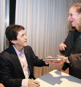 Photo of Mitch Albom, seated at a table, pen in right hand, as he hands a book to a smiling woman.