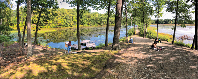 Several students, socially distanced, paint on a tree-lined spit of land jutting into a river on a sunny day.