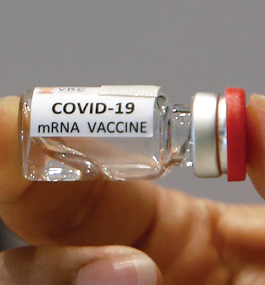 Close-up of a small vial of clear liquid labeled "COVID-19 mRNA Vaccine" held between a thumb and forefinger.