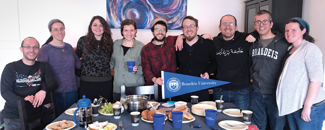 Nine women and men stand behind plates of food on a table with a blue tablecloth. Two of them hold a blue Brandeis pennant.