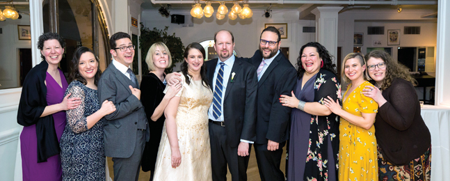 A group of people gather around a woman in a knee-length wedding dress and a man in suit and striped tie.