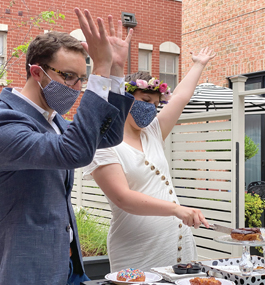 A woman and man, wearing face masks and photographed outdoors, hold their hands in the air in celebration.