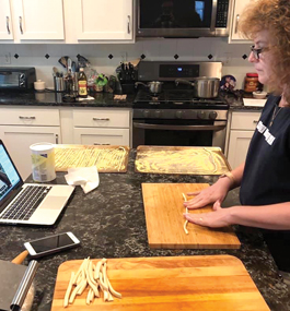 Photo of Denise Silber Brooks standing in her kitchen and looking at her laptop while hand-rolling pasta.