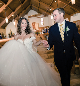 A man and woman in traditional wedding attire walk down the aisle in a chapel with a peaked roof and beams on the ceiling.
