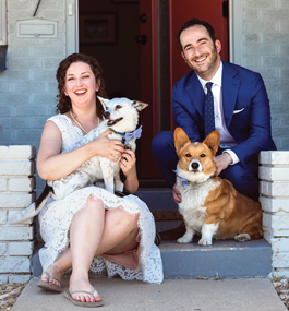 A woman and a man sit on a stoop with two dogs and smile brightly for the camera. The man is in a suit and tie. The woman wears a knee-length wedding dress with flip-flops.