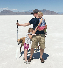 Photo of Eric Harvey holding a cane against a stark white landscape and blue skies, with his two young daughters and a greyhound.