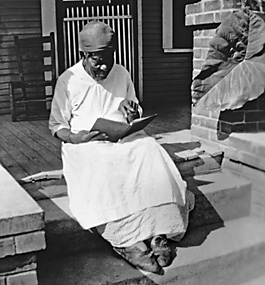 Portrait of former slave Mary Armstrong, wearing a long dress, reading a book while seated on a top step leading to a porch.