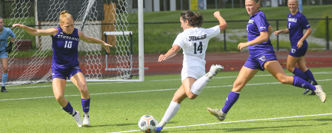 A woman in a blue Stonehill College uniform stretches her arms wide to try to block a shot on net by a woman in a white Brandeis uniform, who has just drawn her foot back to kick.