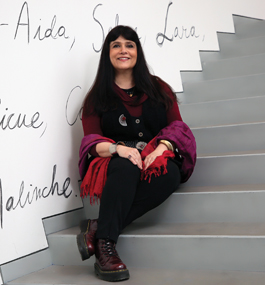 A woman with long dark hair sits on gray steps next to a white wall on which words are written in cursive handwriting