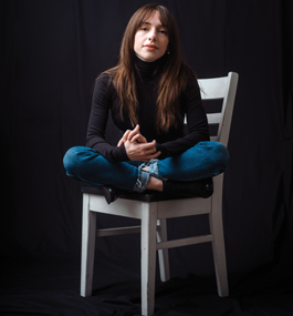 Bruenig, a woman with dark straight hair and bangs, sits cross-legged on a white wooden chair against a dark backdrop.