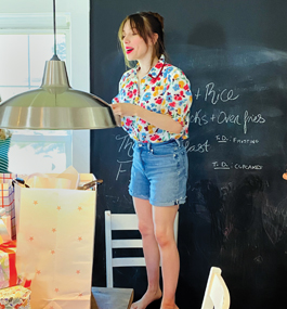 Bruenig stands on a white chair, wearing a brightly patterned shirt and cutoff jeans.