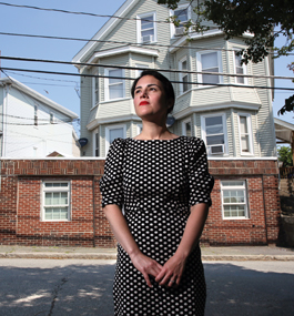 A dark-haired woman in a black-and-white print dress stands in front of two old multistory houses.