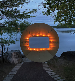 A large round unpainted wooden disk — with a rectangular hole in the middle, surrounded by string lights — overlooks a wooded pond at dusk.