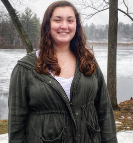 Headshot of a smiling woman in a dark-green hooded jacket, against a backdrop of pond and trees.
