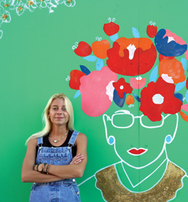 A woman with long blonde hair wearing denim overalls stands with arms crossed in front of a green wall decorated with an outline drawing of Ruth Bader Ginsburg wearing flowers on her head.