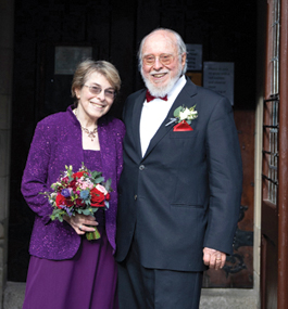 A woman holding a bouquet and a bearded man in a tuxedo stand together in a grand wooden doorway.