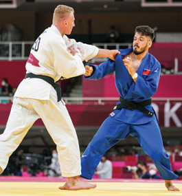 One judo athlete, dressed in a white uniform, grabs the lapel of the blue uniform the other athlete wears, against the dark-red backdrop of a mostly empty stadium.