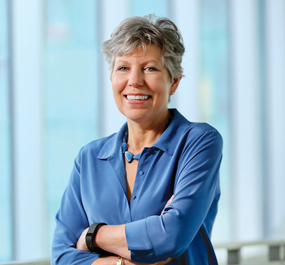 Photo of a woman in a blue shirt standing with her arms crossed in front of a wall of windows.