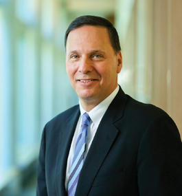 Photo of a smiling man in a dark suit, white shirt and blue striped tie.