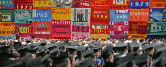 Image from a past Commencement, showing graduates wearing mortarboards (shot from behind), looking toward colorful banners representing Commencement ceremonies from years past.
