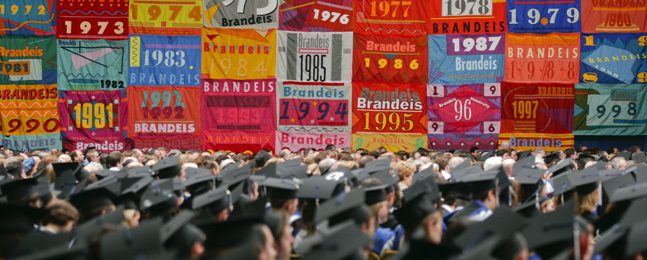 Image from a past Commencement, showing graduates wearing mortarboards (shot from behind), looking toward colorful banners representing Commencement ceremonies from years past.