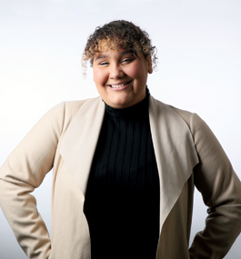 Smiling woman with curly hair, wearing a beige cardigan and dark turtleneck, stands in front of a plain light-colored background.