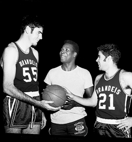 A young K.C. Jones, wearing a plain white T-shirt, holds a basketball while standing between two Brandeis basketball players in their uniforms, who are also touching the ball.