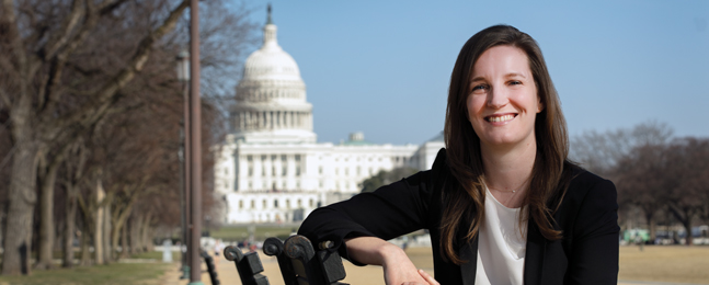 A smiling woman with long brown hair, wearing a white top and a black jacket, sits on a park bench on D.C.'s National Mall with the U.S. Capitol in the background.